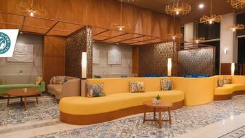 Ein moderner Lounge-Bereich mit einer gebogenen gelben Couch, einer grünen Couch und einem kleinen runden Tisch. Es sind dekorative Paneele und ein gemusterter Boden vorhanden.