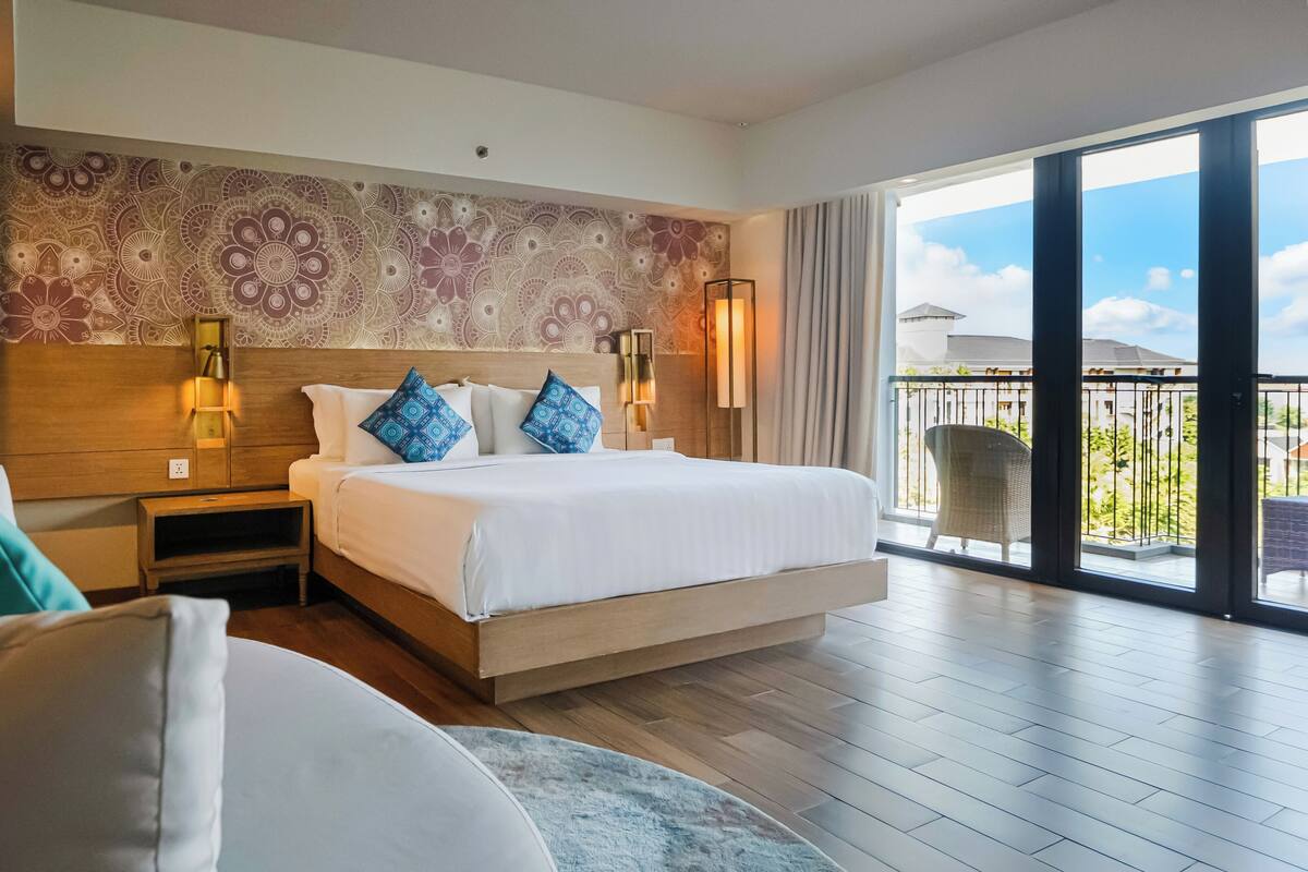 A hotel room with a large bed, a nightstand, a wall with a floral pattern, and a balcony with a view.