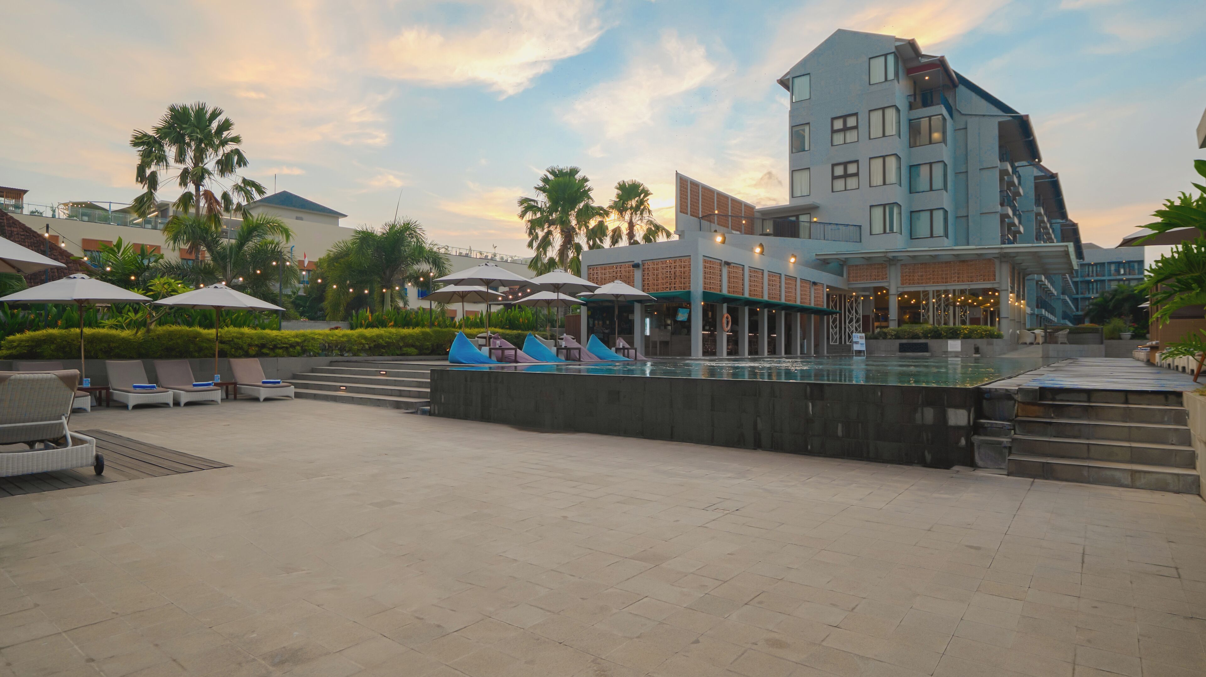 Un patio de hotel con piscina, sillas de descanso, sombrillas y un edificio moderno de varios pisos.
