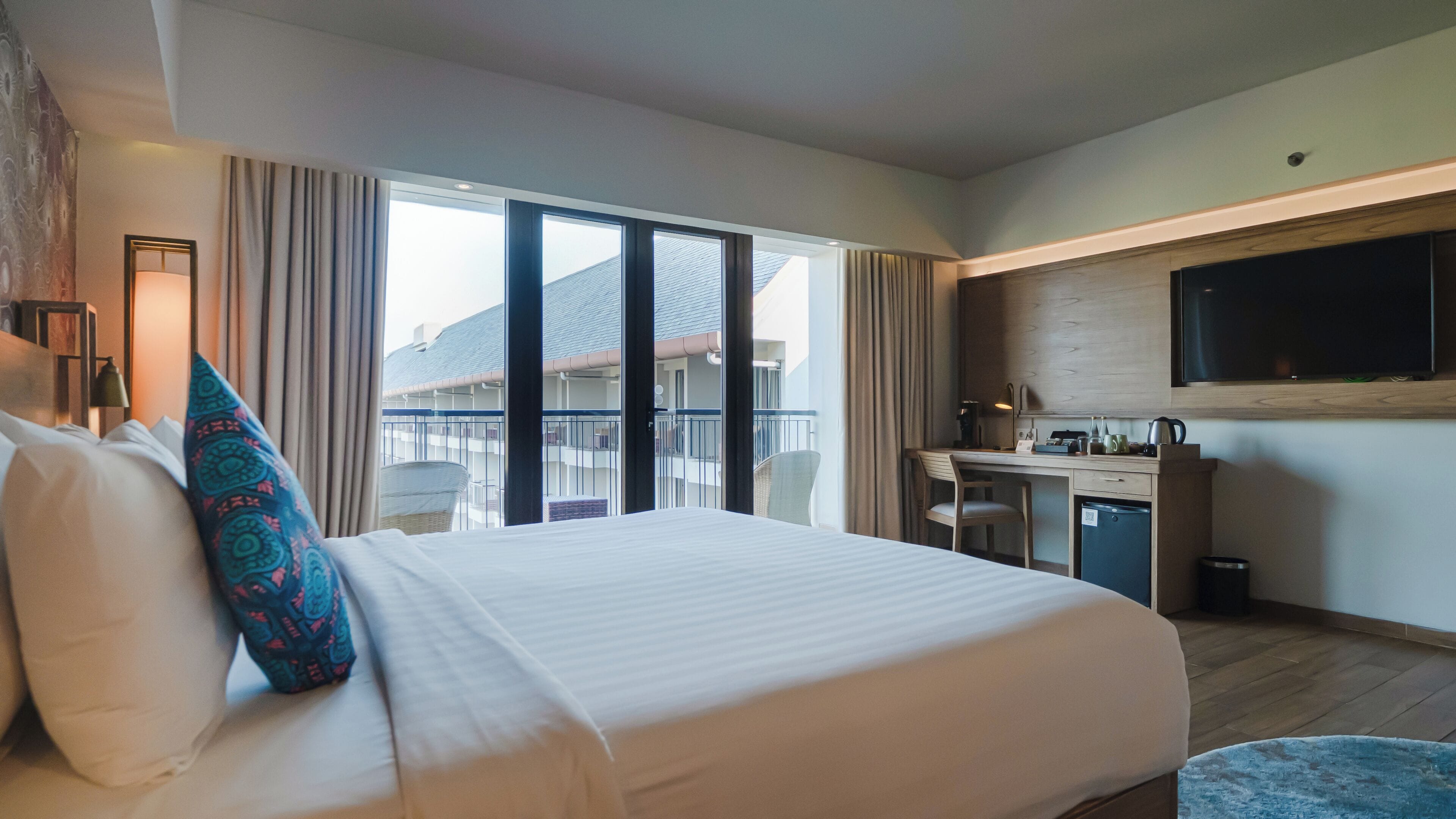 A hotel room with a large bed, a desk, a television, and a balcony with a view of buildings.