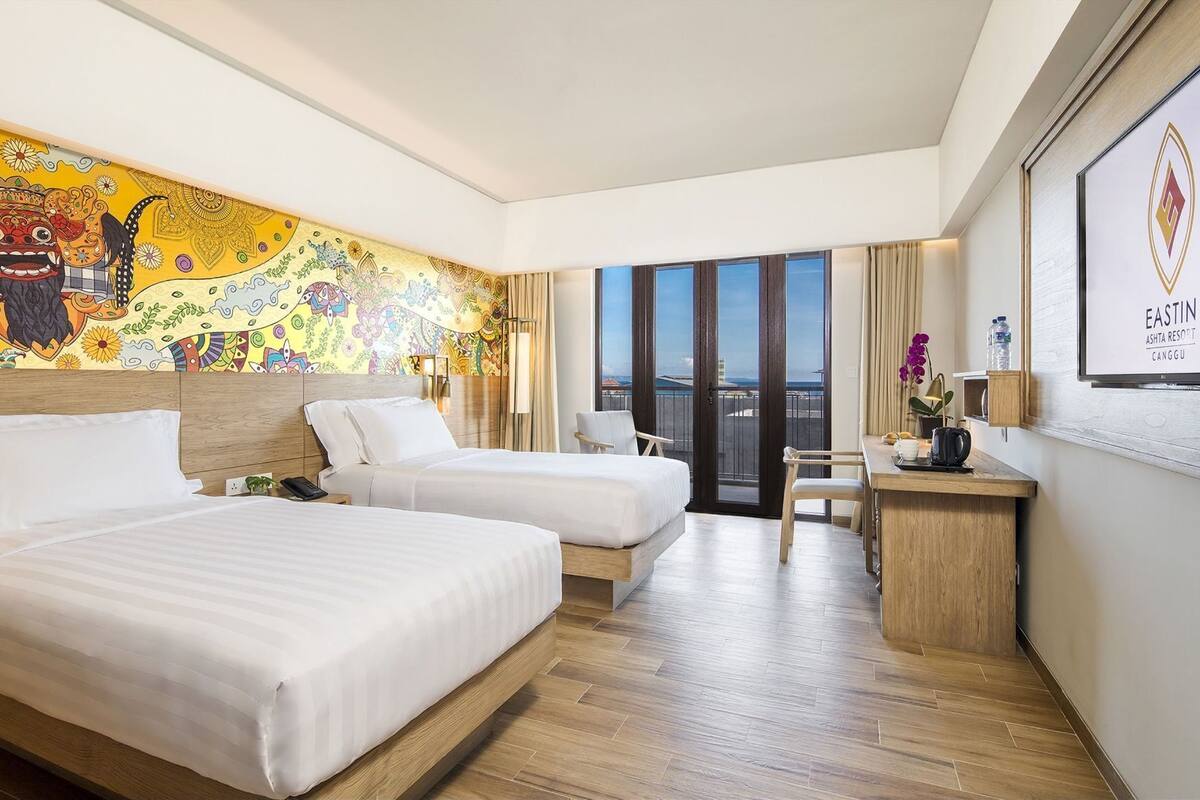 A hotel room with two beds, a desk, a chair, a window with a view, and a colorful mural on the wall.
