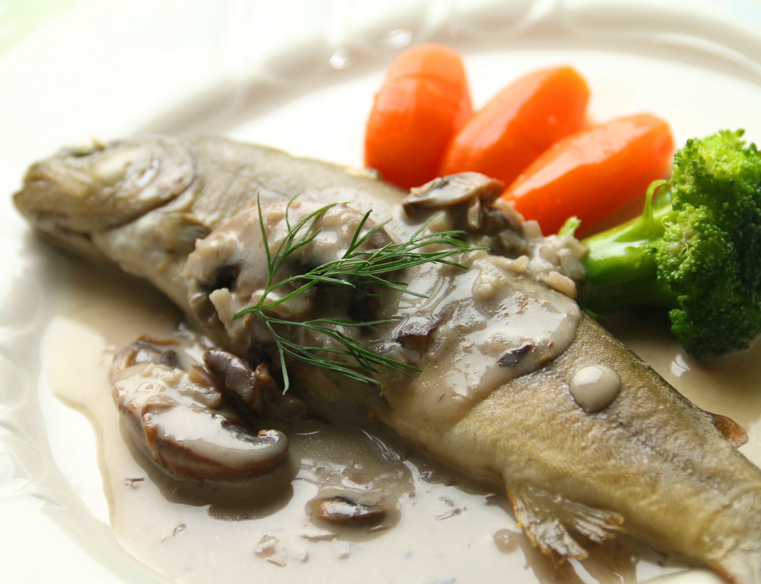 A fish fillet with mushrooms and dill on a plate, accompanied by carrots and broccoli.