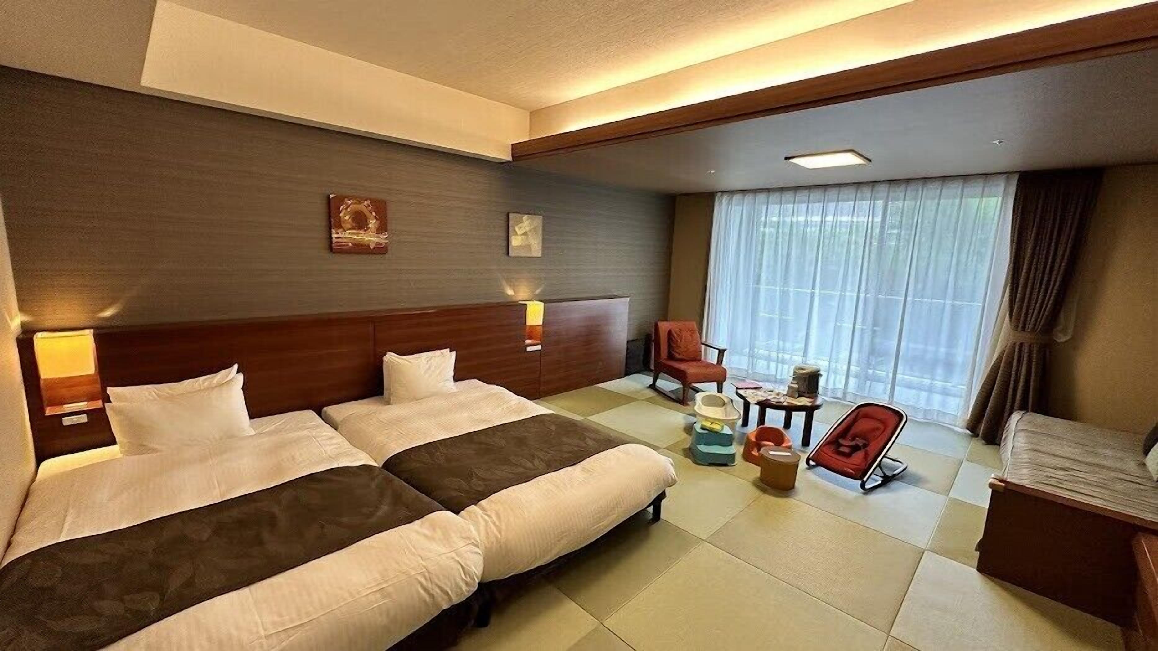 Hotelkamer met een groot bed, een kleiner bed, een bureau, een stoel en een raam met gordijnen.