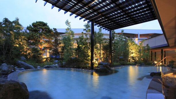 A serene outdoor pool area with a wooden pergola, surrounded by rocks and trees, and a building in the background. - Hotel WELLSEASON Hamanako (Hamamatsu)