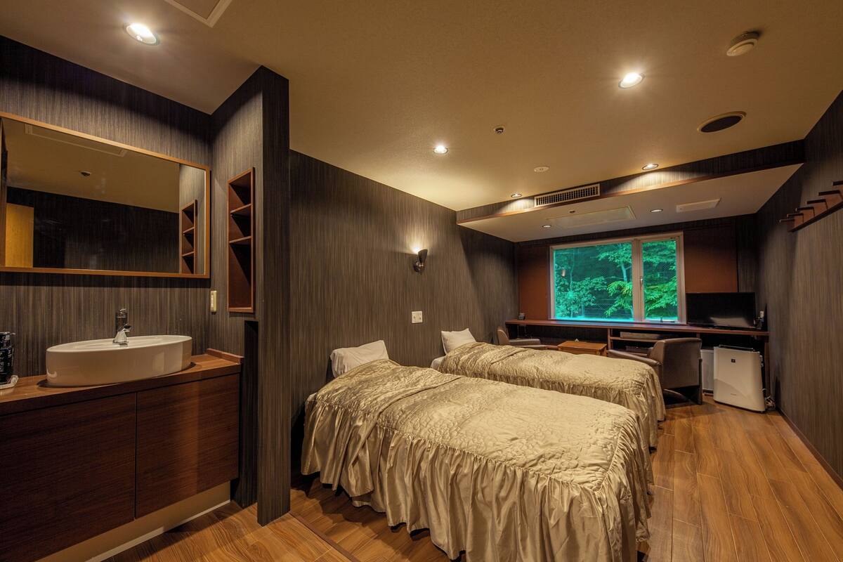 A modern hotel room with two beds, a large mirror, a vanity with a sink, and a desk with a computer.