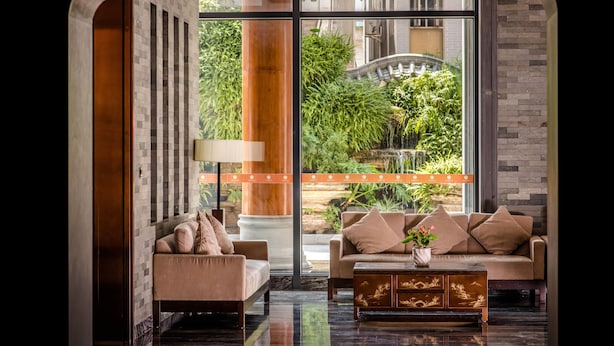 A modern living room with a sofa, armchair, and coffee table, large windows with a view of greenery and a bridge, and a decorative lamp. - Xiang Yun Sha Garden Hotel (Foshan)