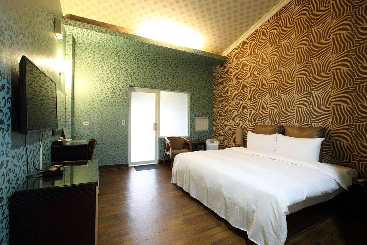 A hotel room with a bed, a desk with a chair, a television, and a patterned wall.