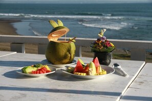 A table with plates of fruit and a coconut drink on a beach overlooking the ocean. - PNB Beach Resort (Pererenan)