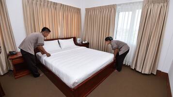 A neatly made bed in a hotel room with a wooden headboard, a bedside table with a lamp, and beige curtains.
