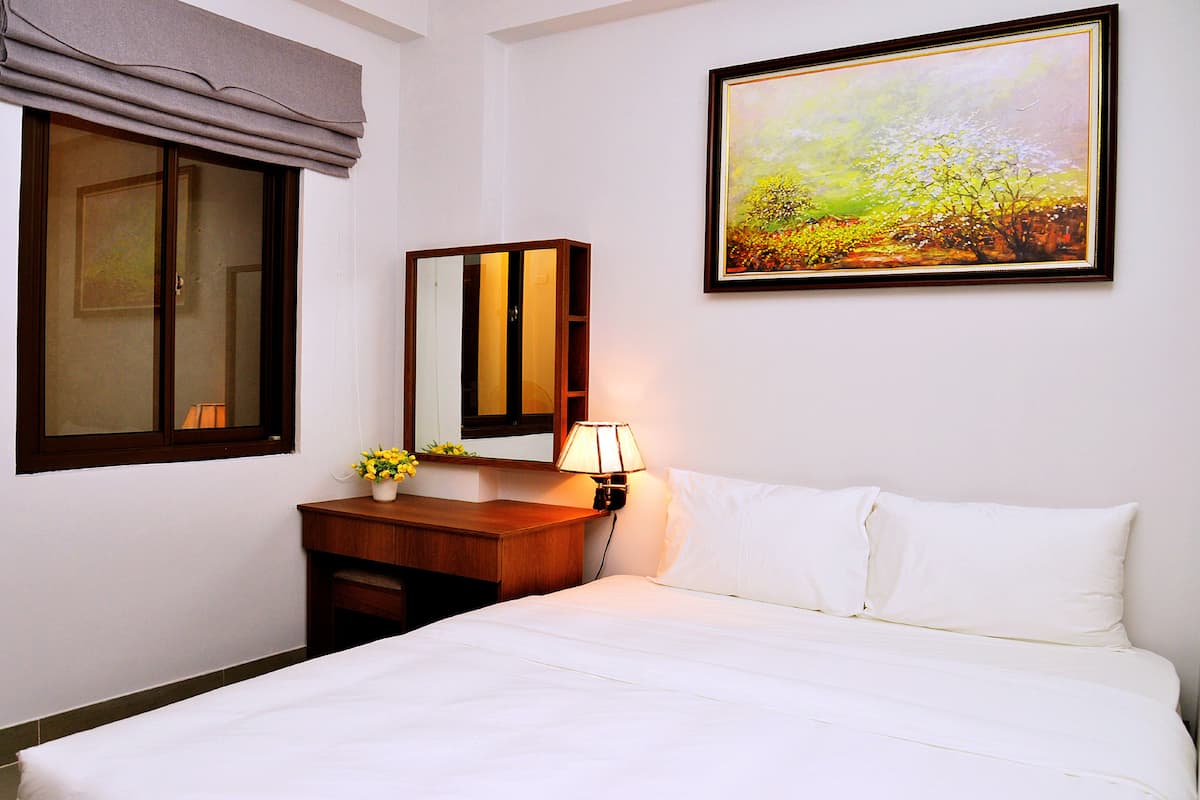 Une chambre d’hôtel avec un lit, une table de chevet avec une lampe, un miroir, une fenêtre avec un store et un tableau au mur.