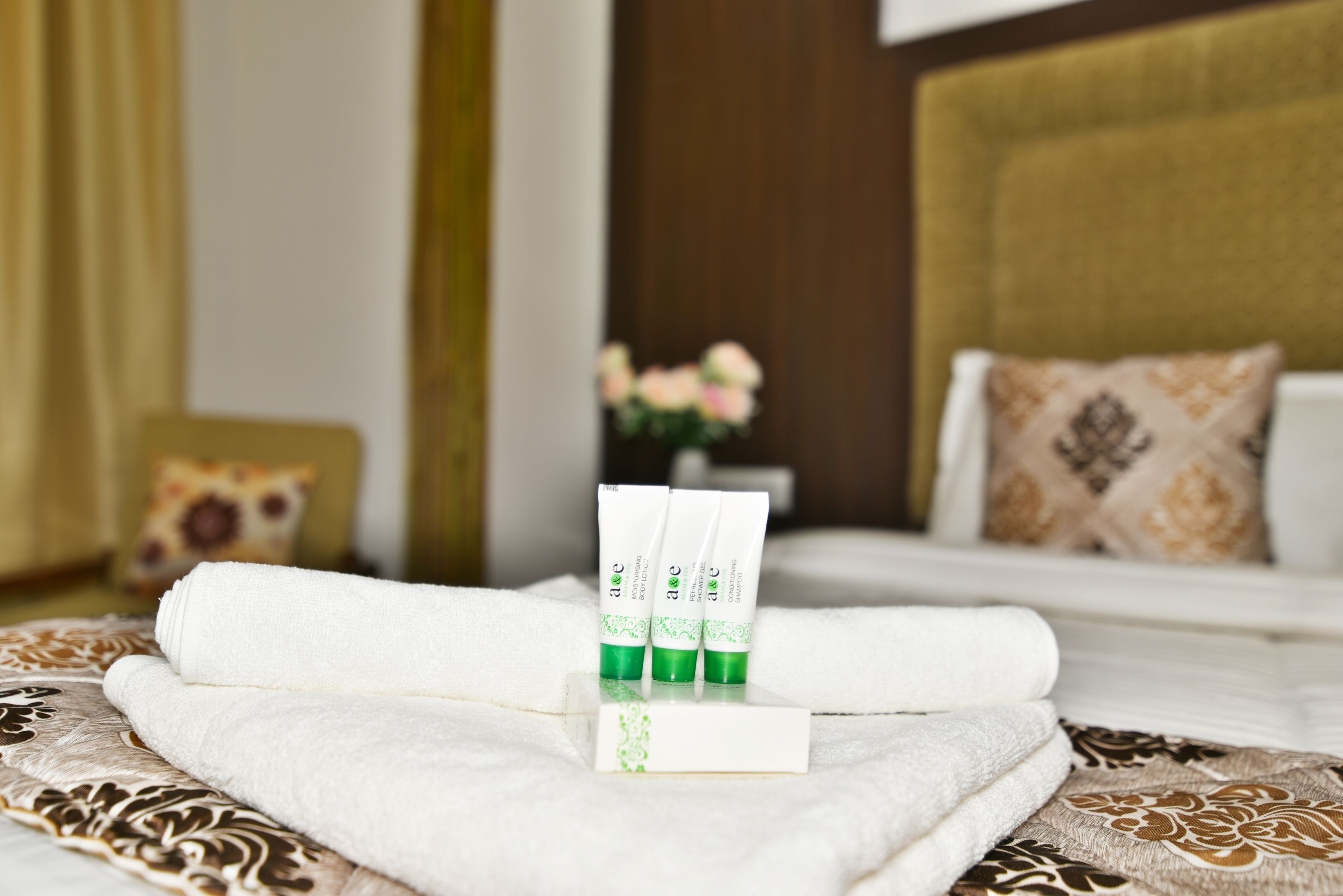A neatly made bed with a white towel and three tubes of aloe vera gel on a patterned bedspread.