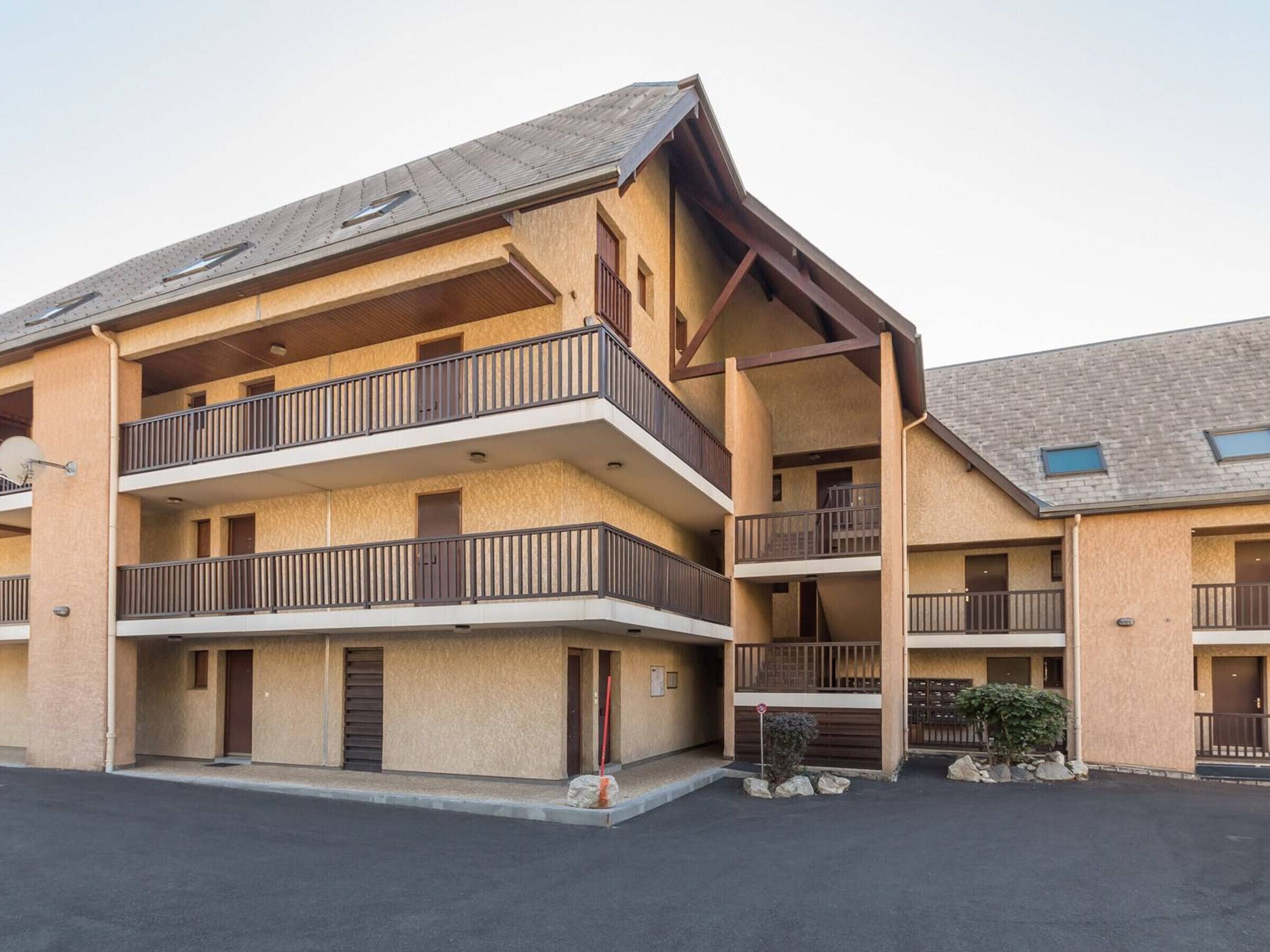 A multi-story building with balconies, a flat roof, and a parking area in front.