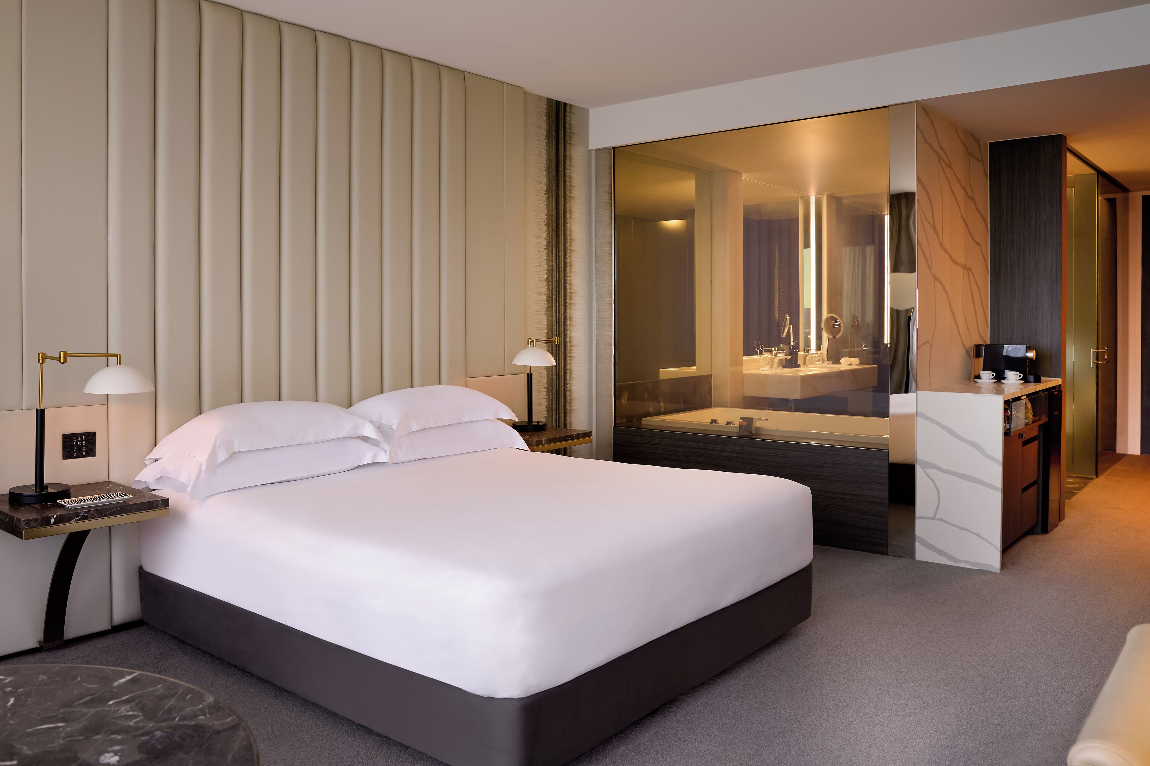 A modern hotel room with a large bed, bedside tables with lamps, a bathroom with a bathtub, and a desk with a coffee maker.