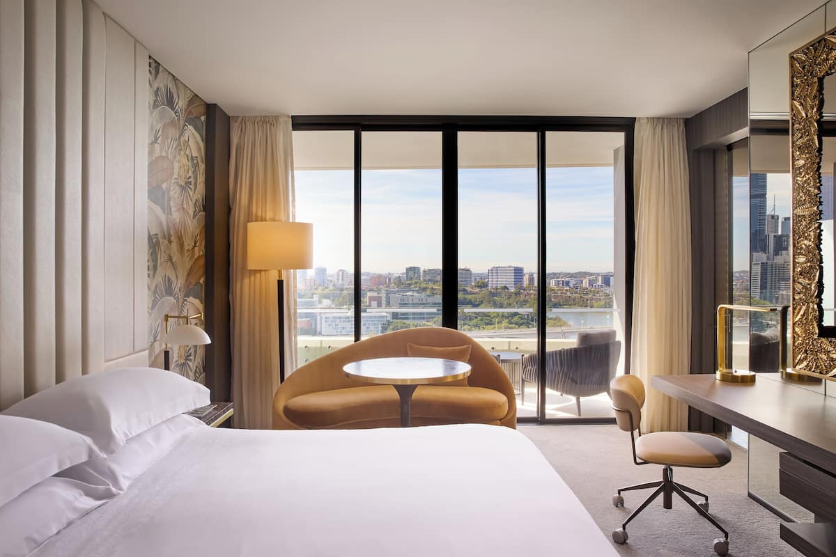 Een hotelkamer met een groot bed, een ronde tafel, een bureau en een stoel. De kamer heeft een groot raam met uitzicht op de stad.