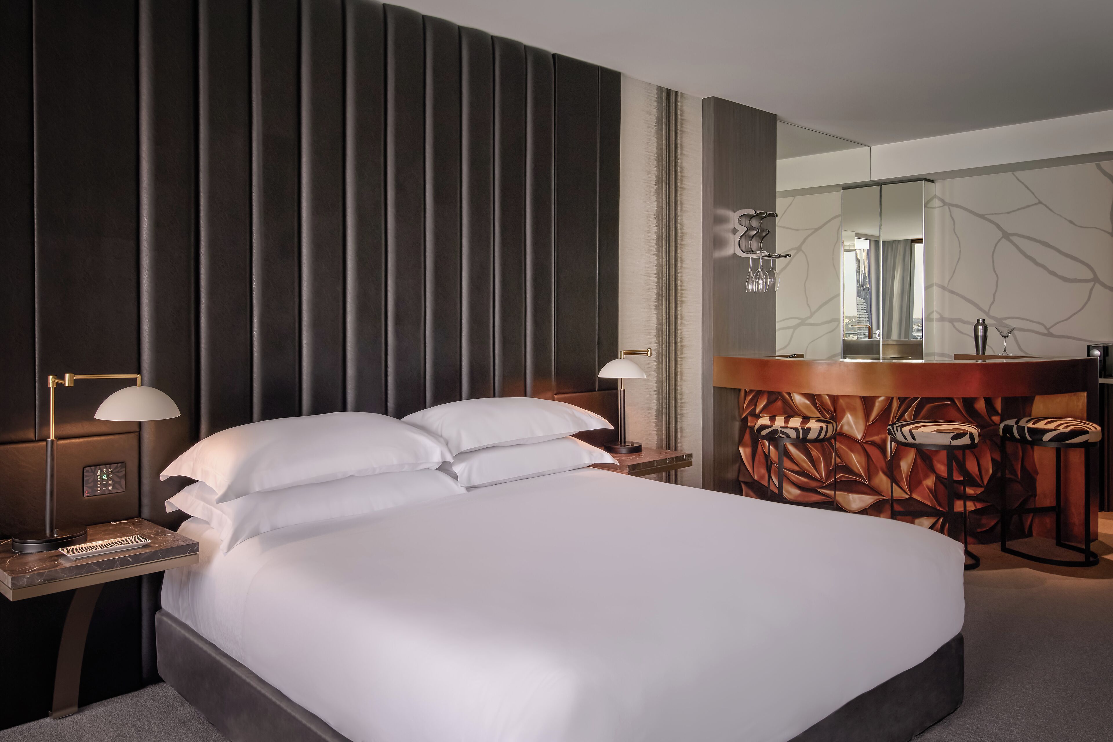 A modern hotel room with a large bed, bedside lamps, a vanity with a mirror, and a seating area with stools.