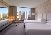 A hotel room with two beds, a desk with a chair, a lamp, and a view of the cityscape.