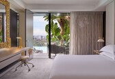 A hotel room with a large bed, a desk with a chair, a view of a cityscape and palm trees through a sliding glass door.
