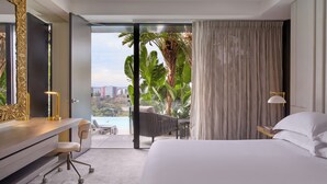 Habitación de hotel con una cama grande, un escritorio con silla, y vistas a la ciudad y palmeras a través de un ventanal corredero.