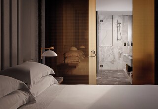 A modern hotel room with a bed, a nightstand, a lamp, and a view into a bathroom with a shower and marble walls.