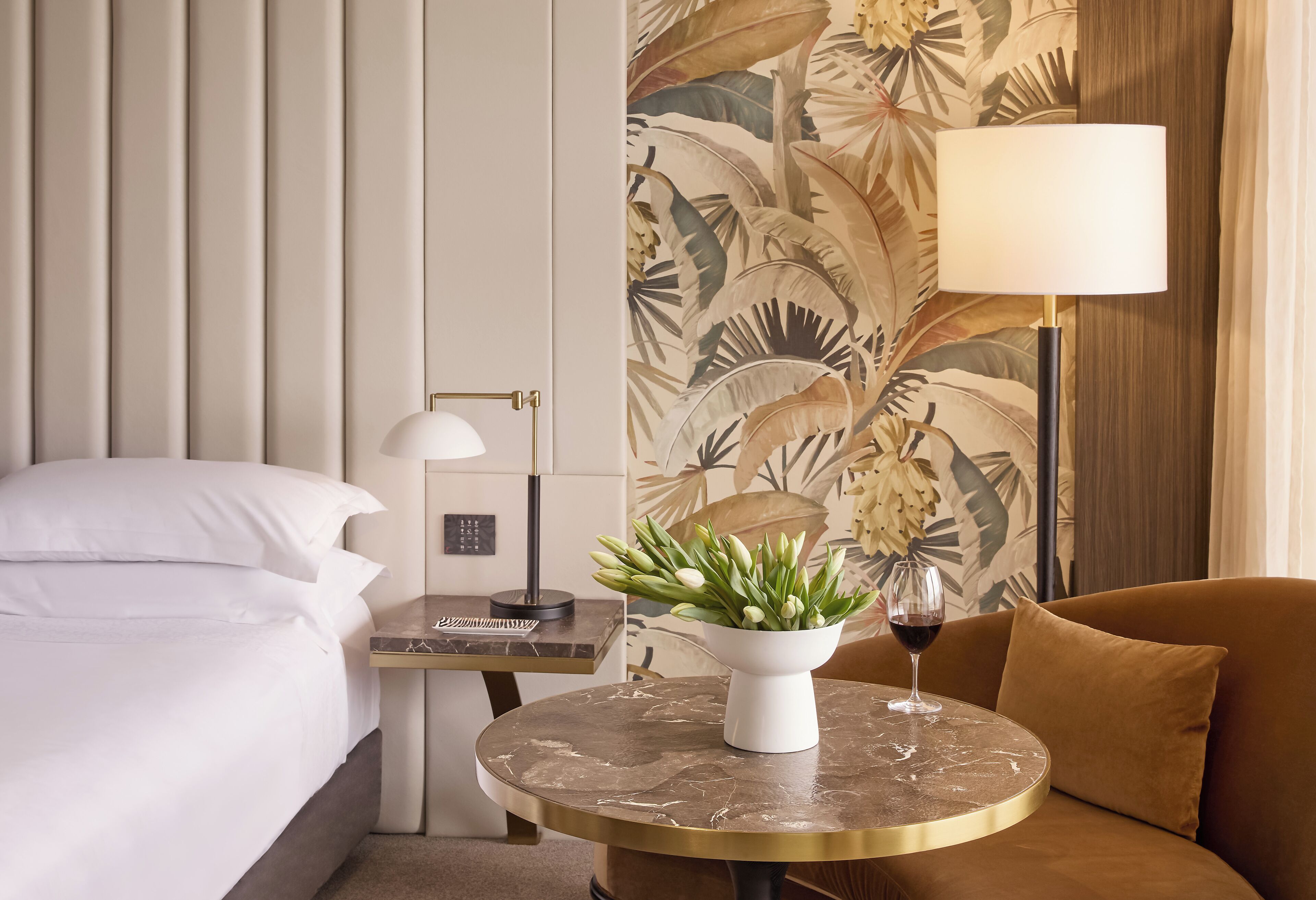 A hotel room with a bed, a round table with a vase of flowers, a lamp, and a wine glass.