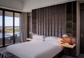 A modern hotel room with a large bed, two bedside tables, a view of a cityscape and a body of water, and a balcony with a chair.
