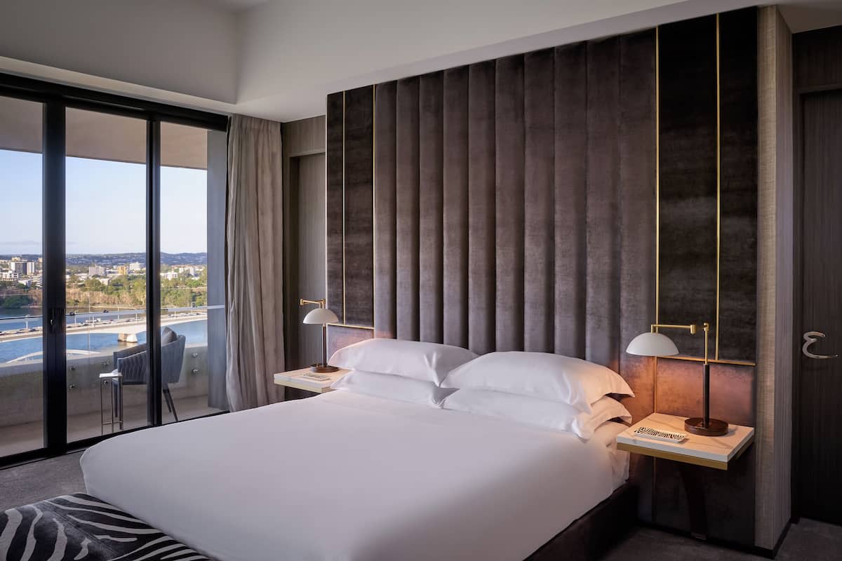 Een moderne hotelkamer met een groot bed, twee nachtkastjes, uitzicht op een stadslandschap en een wateroppervlak, en een balkon met een stoel.