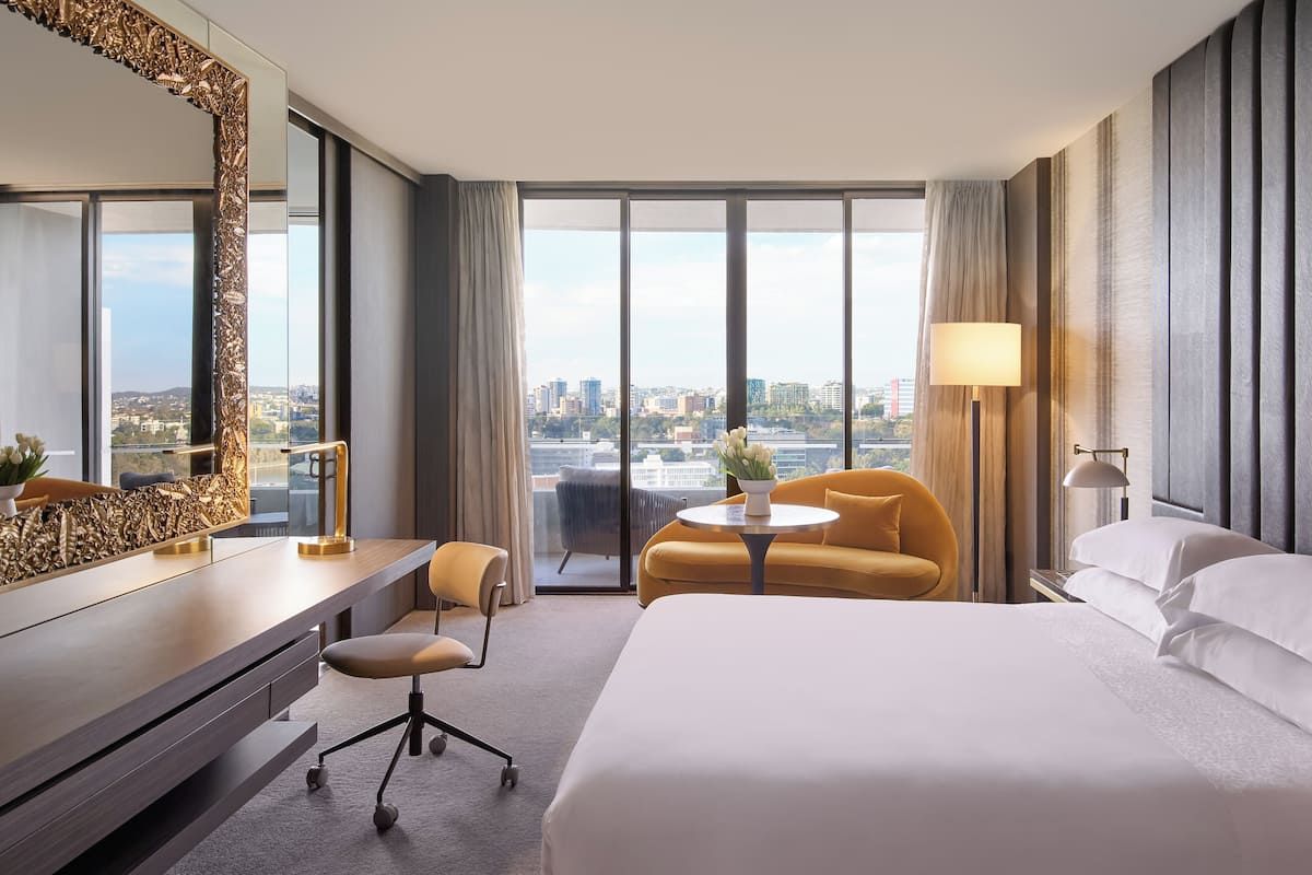 Hotelkamer met een groot bed, een bureau met stoel, een bijzettafeltje met lamp en uitzicht op de stad door grote ramen.