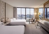 A hotel room with two beds, a desk, a chair, and a large window offering a city view.
