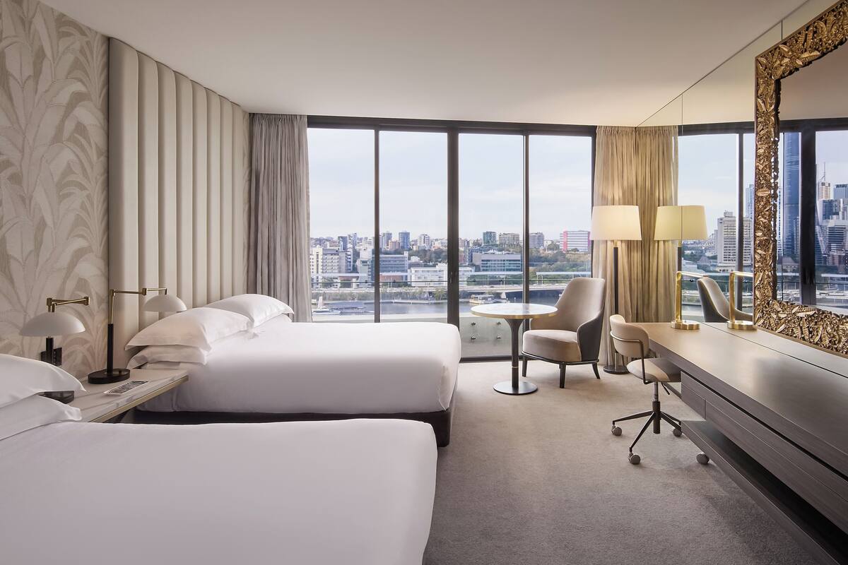 Een hotelkamer met twee bedden, een bureau, een stoel en een groot raam met uitzicht op de stad.