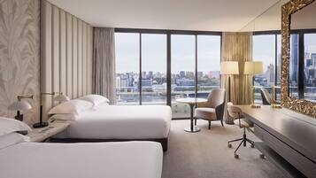 A hotel room with two beds, a desk, a chair, and a large window offering a city view.