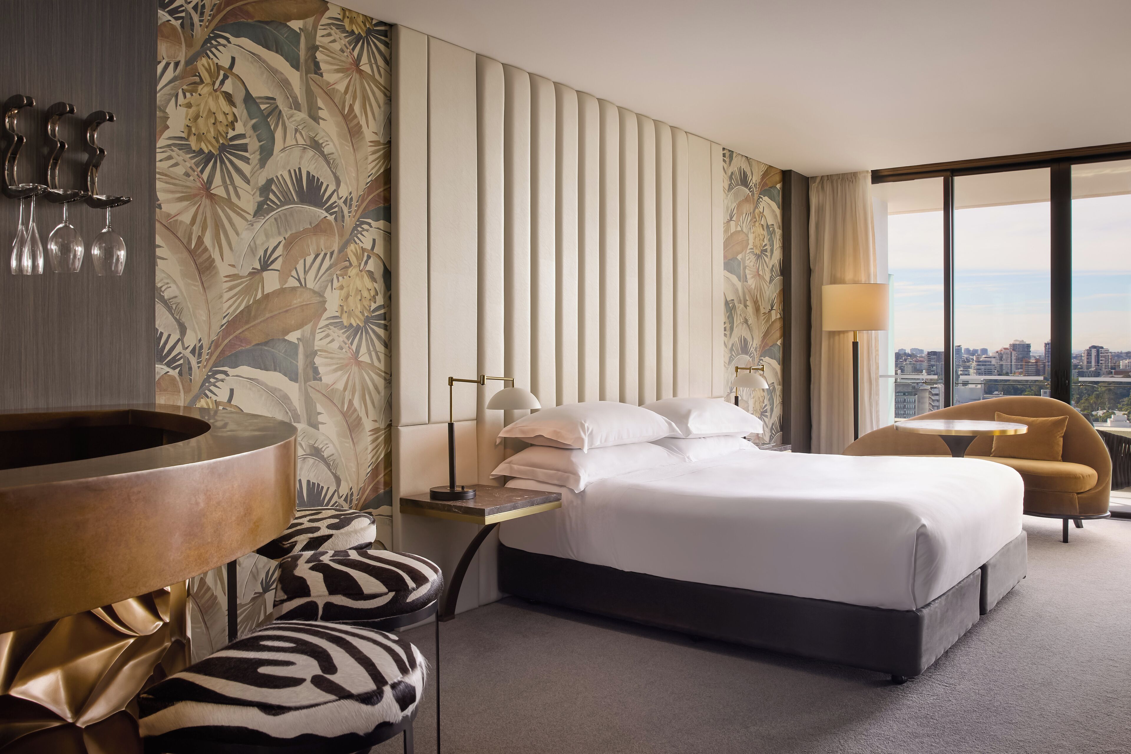 A modern hotel room with a large bed, a sitting area with a chair, a small table, and a large window offering a city view.