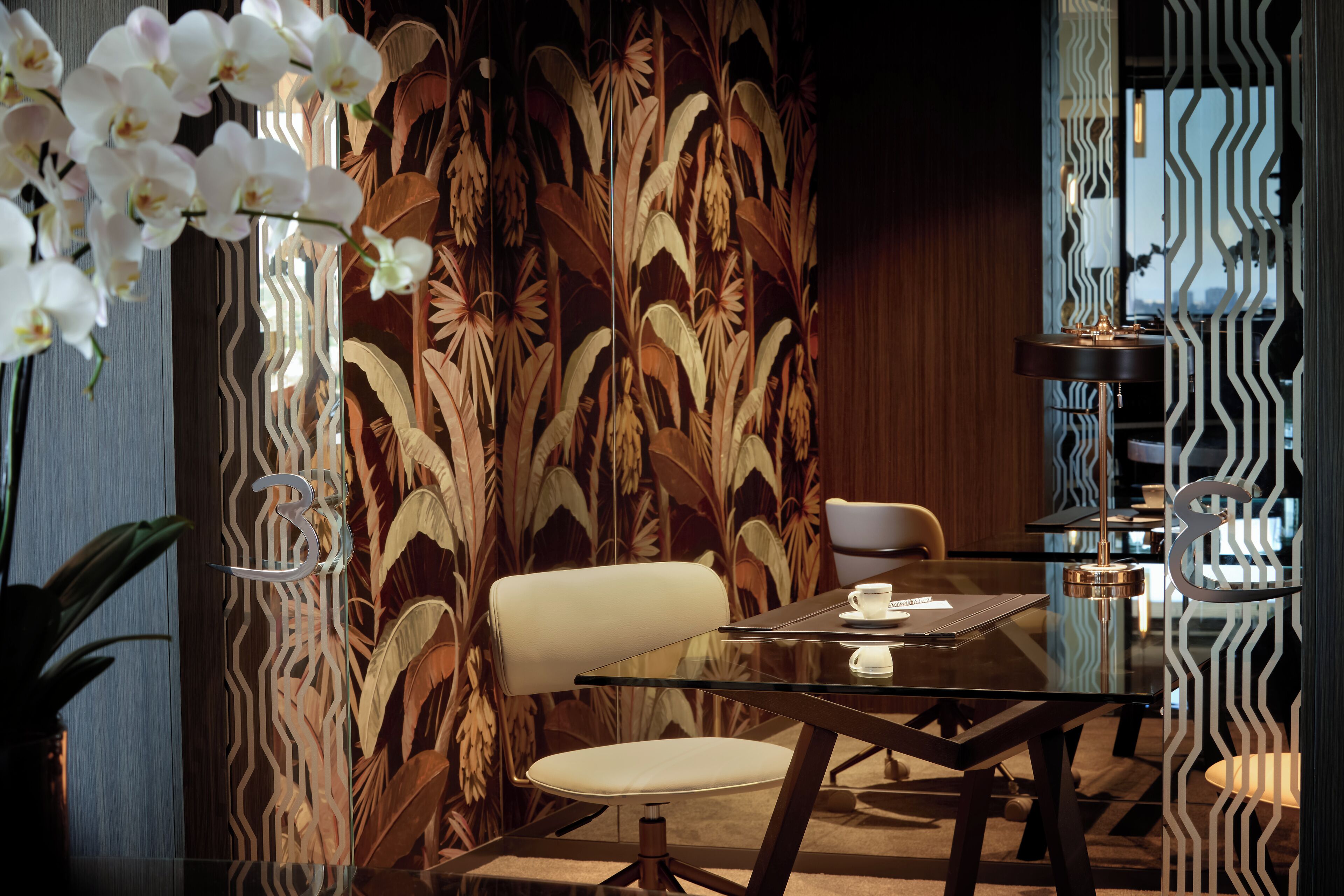 Un comedor moderno con una mesa de vidrio, sillas blancas y un muro decorativo con un diseño de hojas.