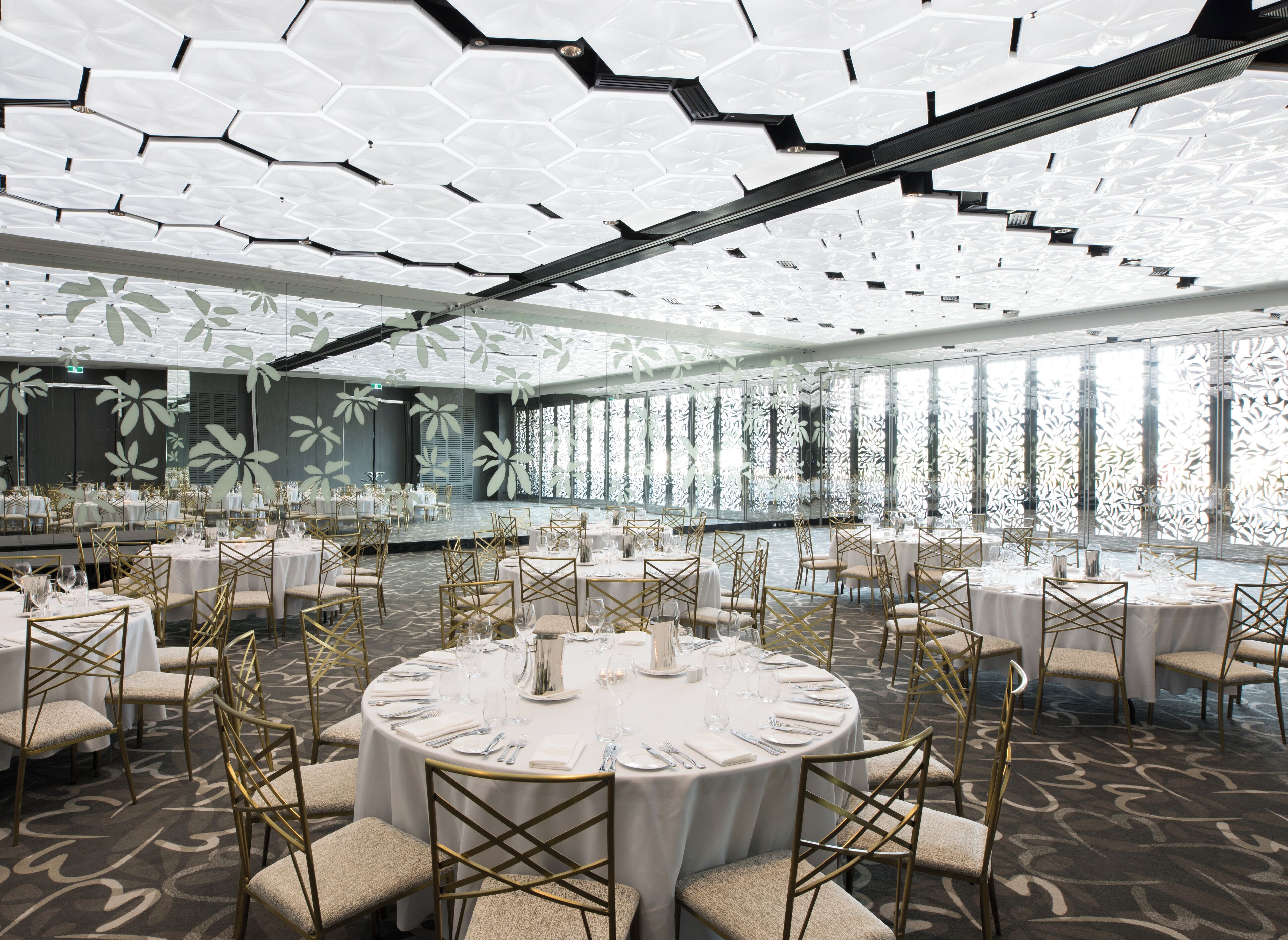 A spacious banquet hall with round tables set for dining, featuring gold chairs and white tablecloths.