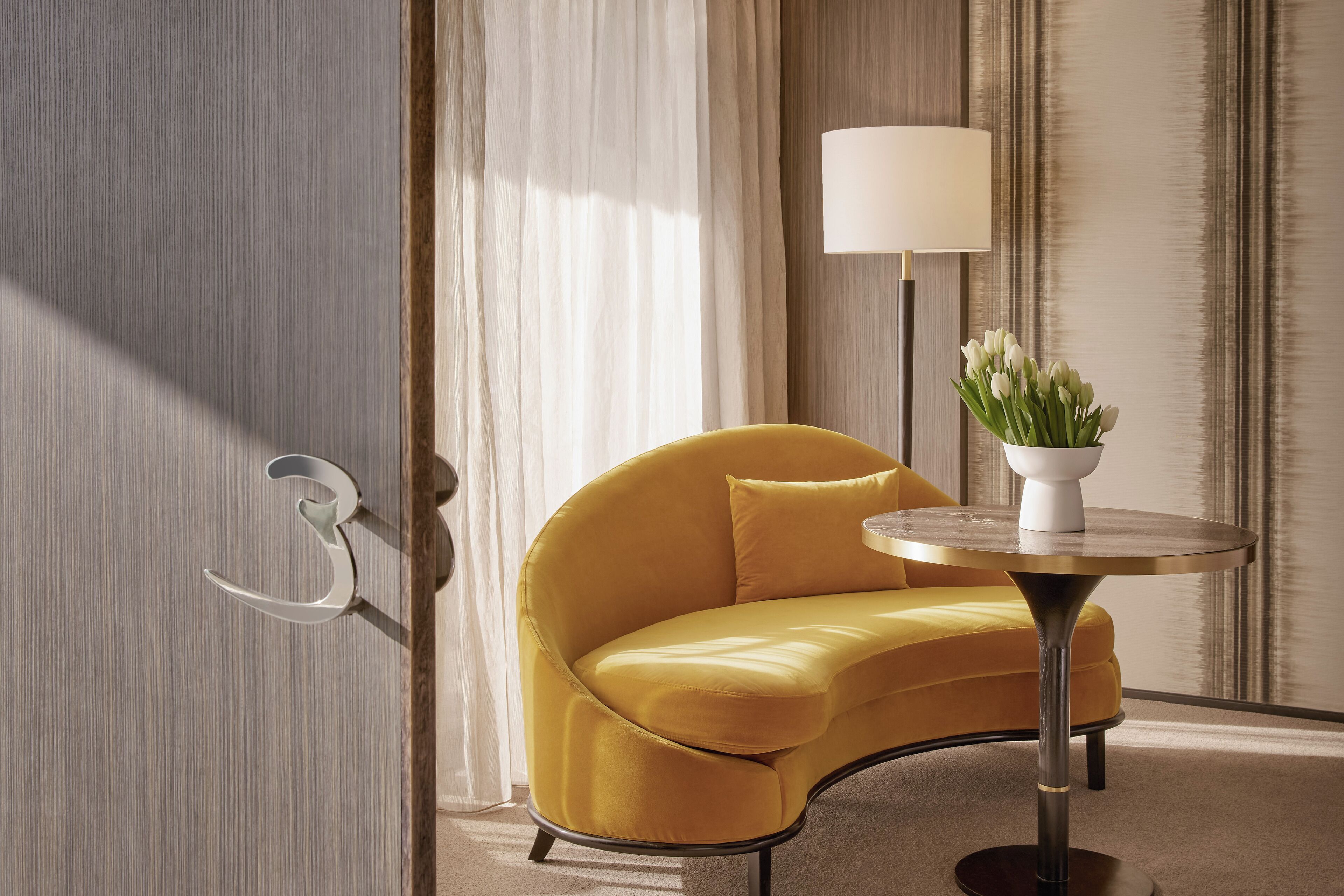 A modern room with a curved yellow sofa, a round table with a lamp, and a vase of white flowers.