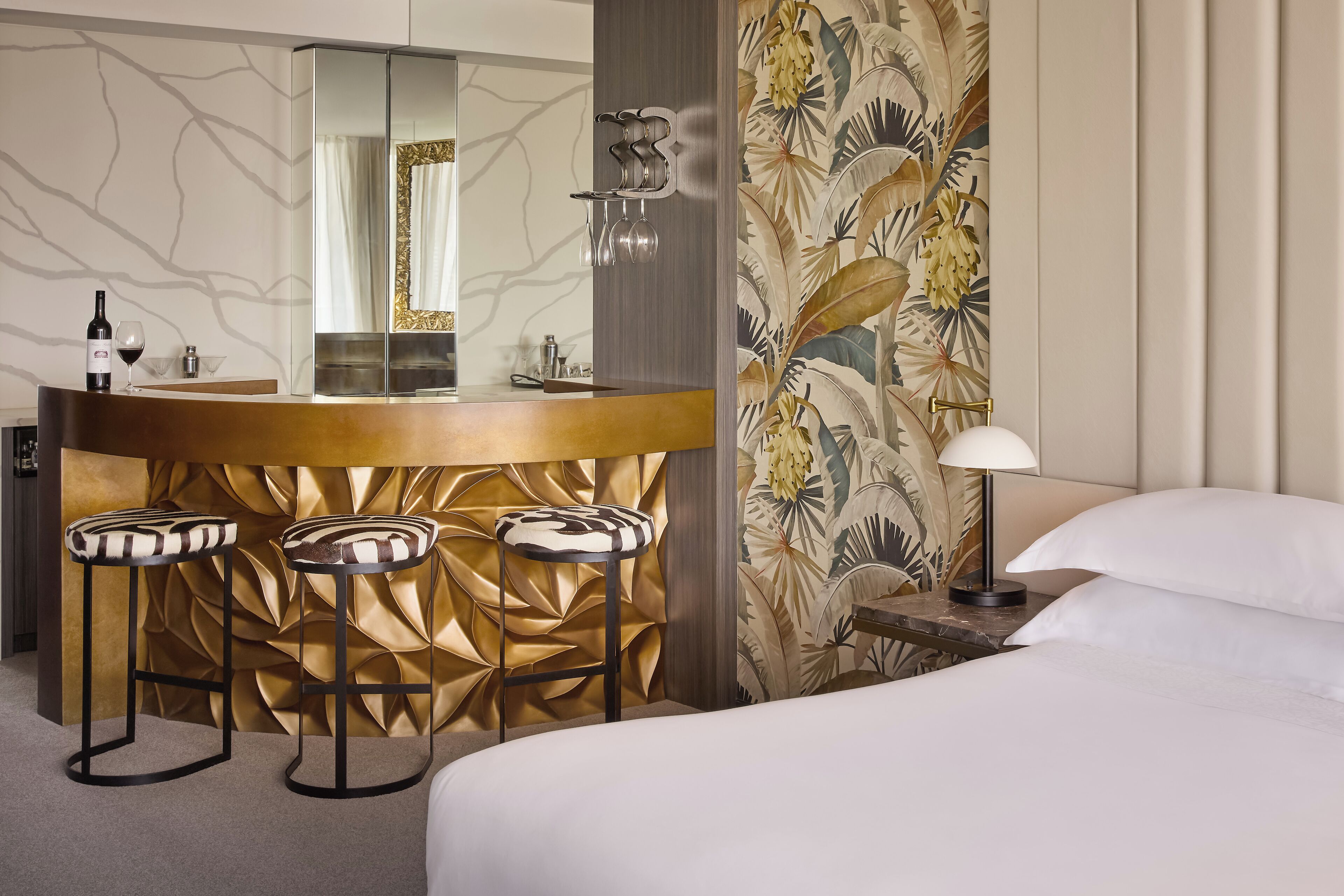 A hotel room with a bed, a bedside table with a lamp, a mirror, a bar area with stools, and a decorative wall.