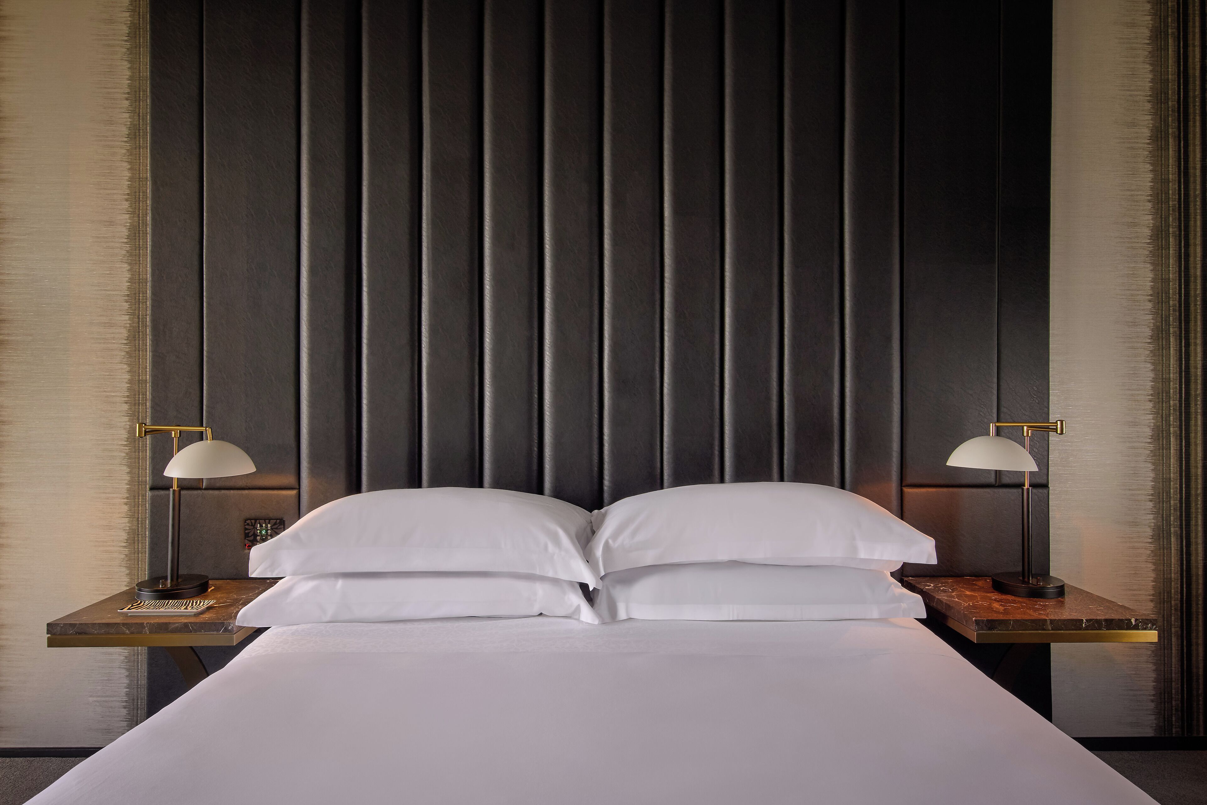 Una camera d'albergo con un letto grande, due comodini con lampade e una testiera scura.