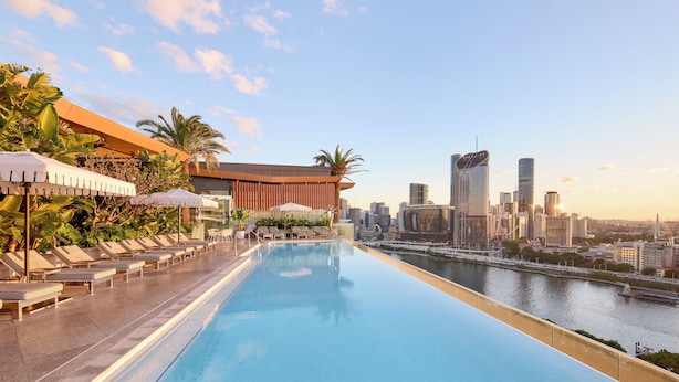 Una piscina infinita en la azotea con sillones de descanso y sombrillas, con vistas a un horizonte urbano y un río.
