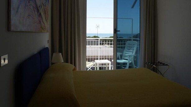 Habitación de hotel con cama, un cuadro en la pared, vista del balcón y del exterior, y una silla en el balcón.