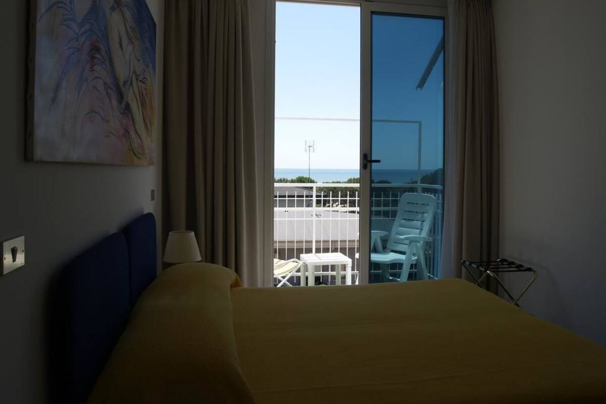 Habitación de hotel con cama, un cuadro en la pared, vista del balcón y del exterior, y una silla en el balcón.