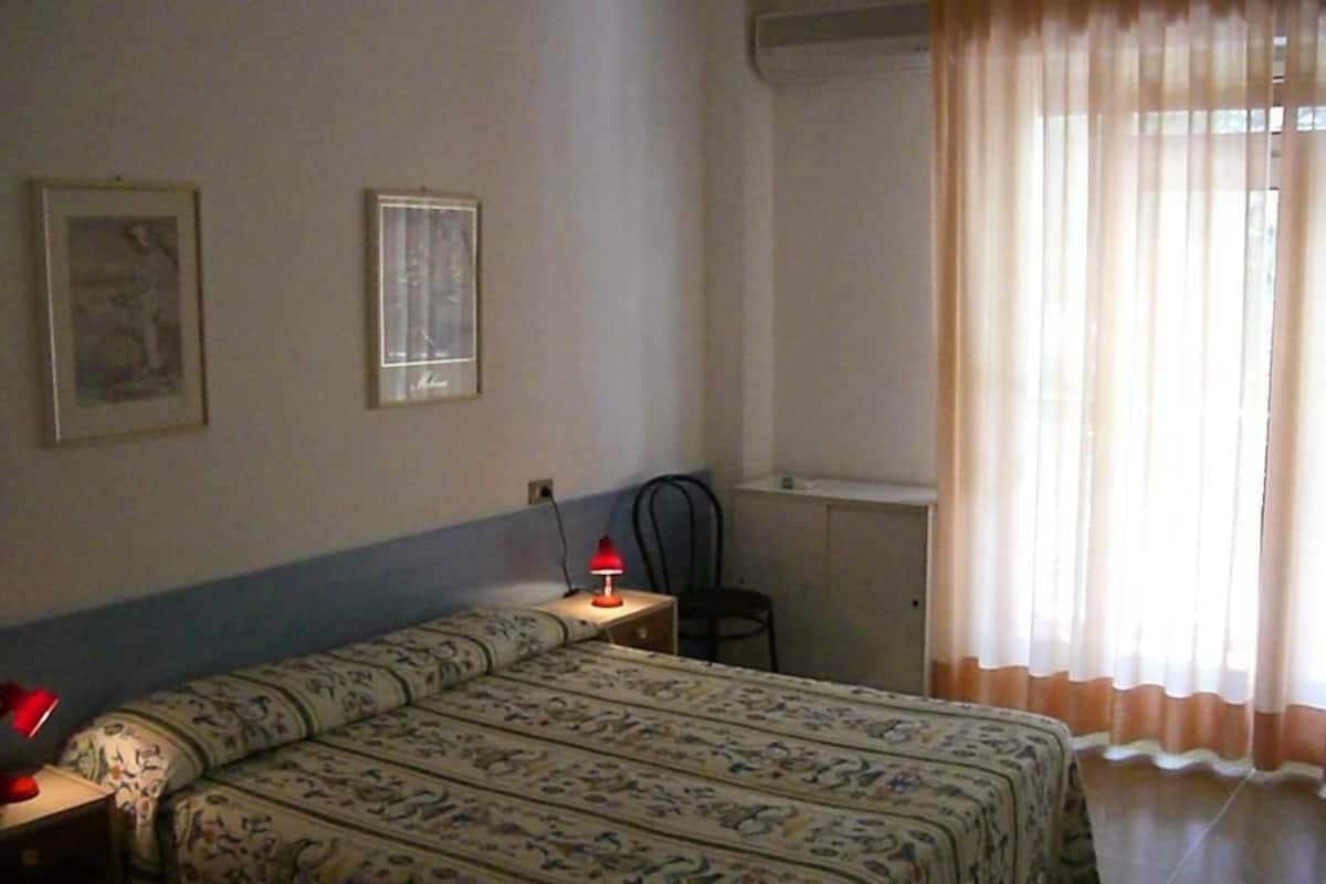 Un dormitorio con cama, mesita de noche, lámpara, silla, escritorio y ventana con cortinas.