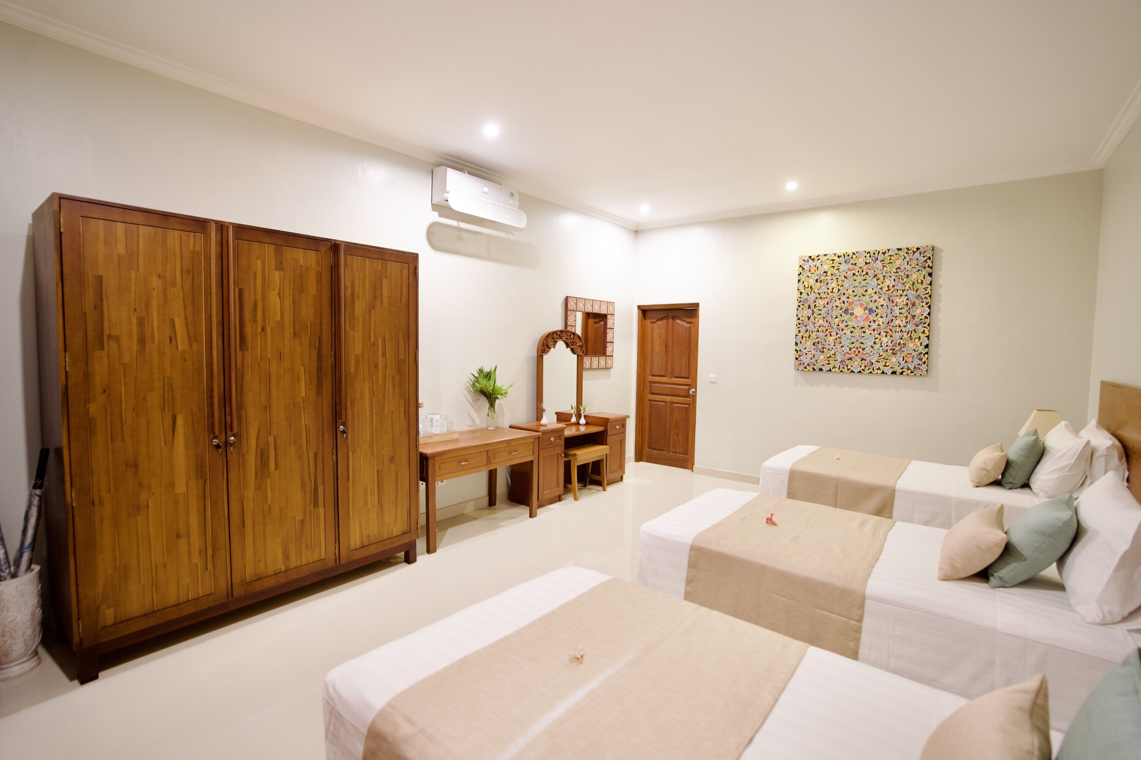 A hotel room with a wardrobe, a desk, a mirror, a bed with a beige bedspread, and a colorful abstract painting on the wall.