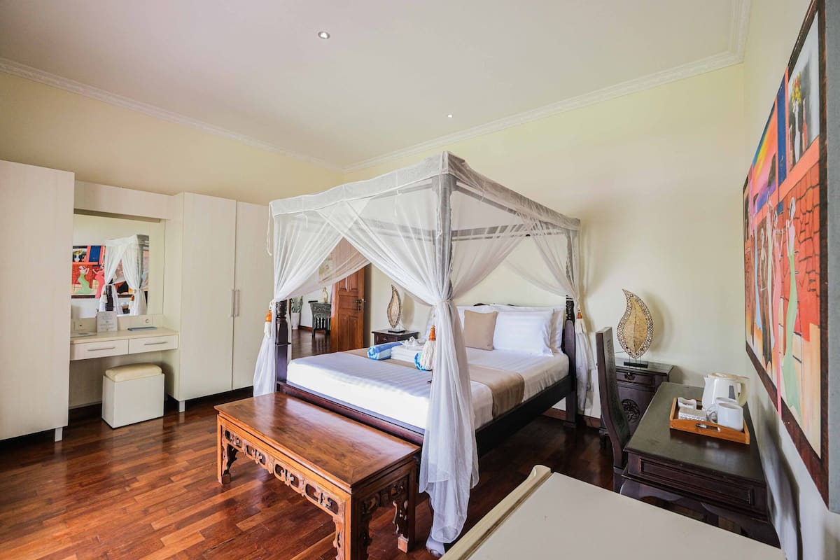 A bedroom with a four-poster bed, a wooden bench, a desk with a chair, and a bedside table with a lamp.