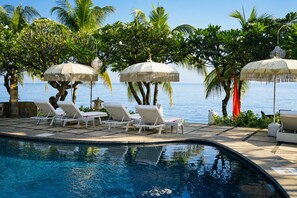 A poolside area with lounge chairs and umbrellas, a clear view of the ocean, and a tropical setting with palm trees.