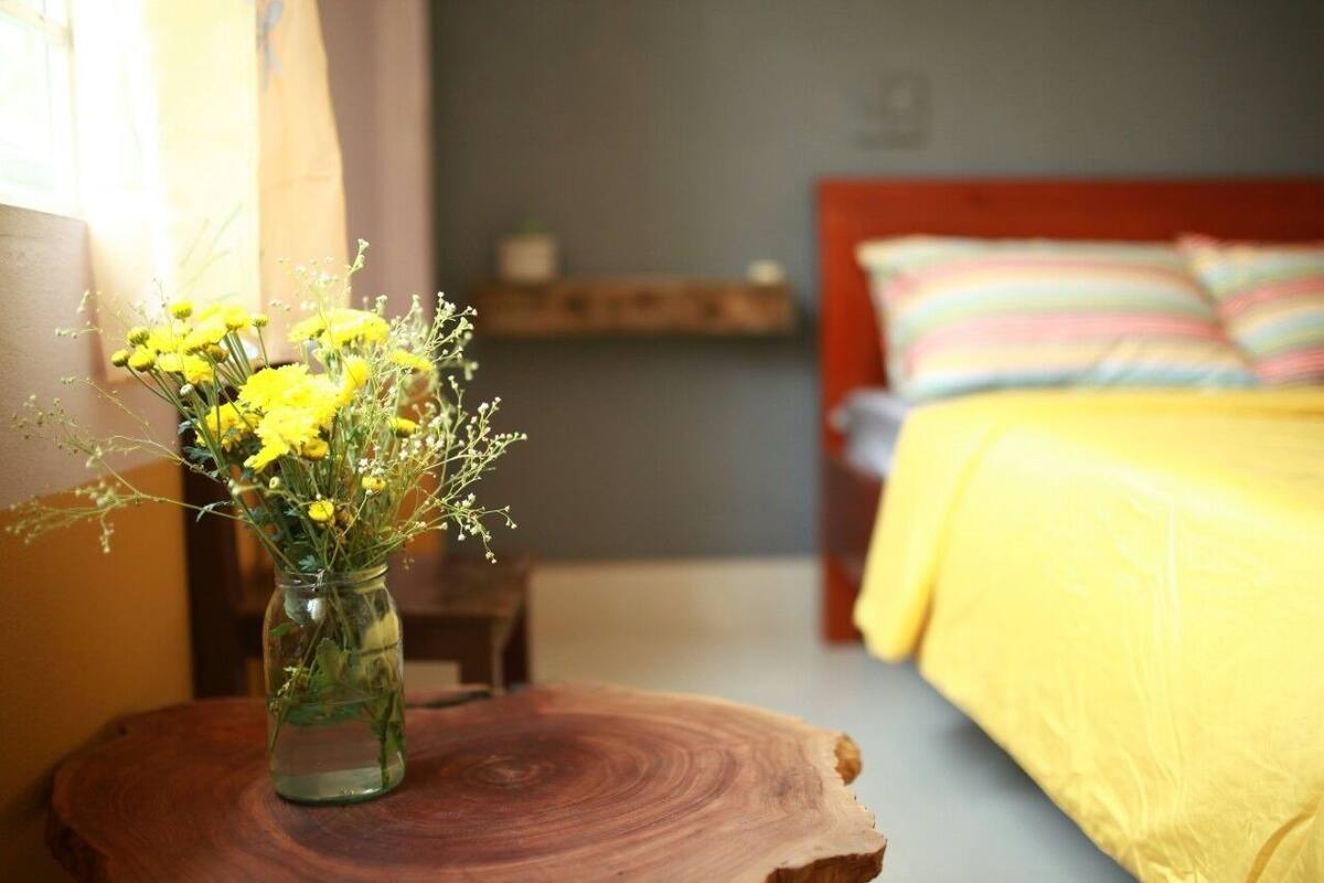 A bedroom with a bed, a bedside table, and a vase with yellow flowers on a wooden stool.