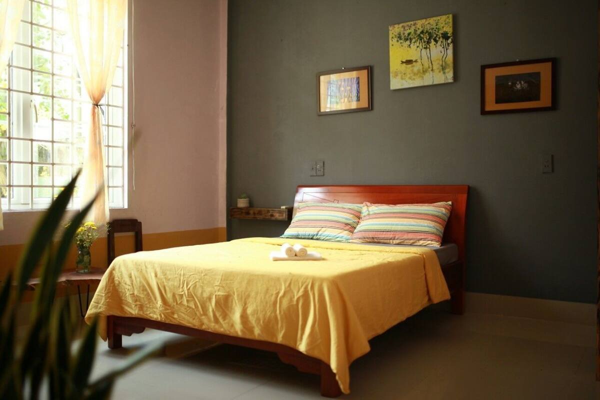 A neatly made bed with a yellow bedspread and striped pillows, a small bedside table with a vase of flowers, and a window with curtains.