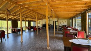 A spacious indoor area with wooden beams, tables, chairs, and a large window providing natural light. - Pinar Kir Evi (Arhavi)