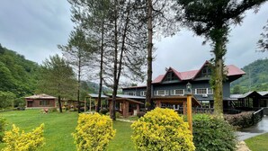 A large, two-story building with a red roof and multiple windows, surrounded by lush greenery and trees. - Pinar Kir Evi (Arhavi)