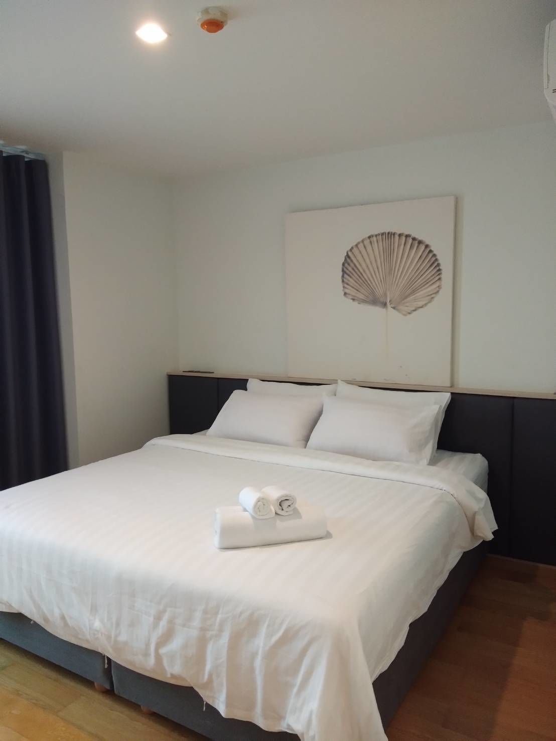 A neatly made bed with white linens and rolled towels, a dark headboard, and a wall-mounted decorative piece.