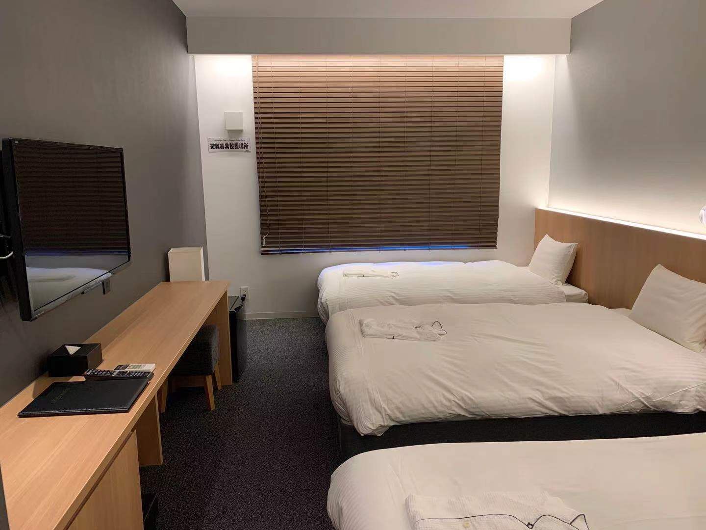 A hotel room with a desk, two beds, a television, and a window with blinds.