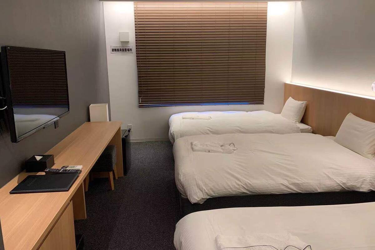 A hotel room with two beds, a desk with a chair, a TV, and a window with blinds.